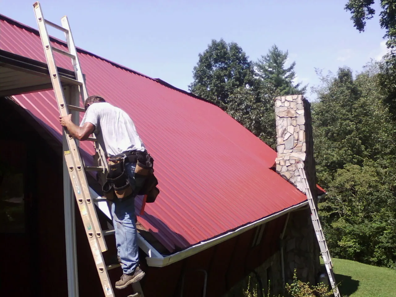 Roofer on a ladder beside a red metal roof and stone chimney — Soffit & Fascia Repair by Roofing Brothers in Branson